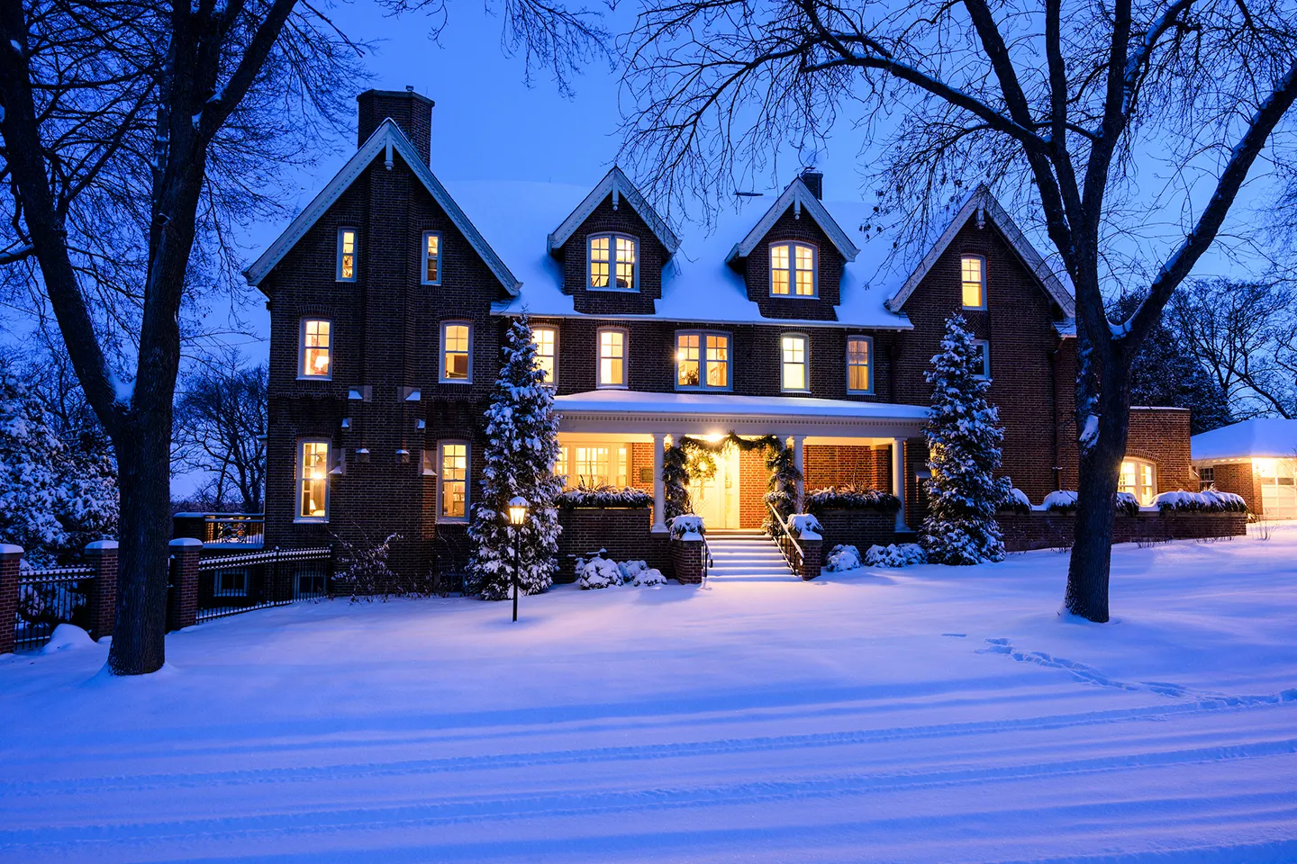 Olin House at dusk, covered in snow, with warm yellow light glowing from the windows.